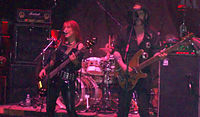 Female musician Enid Williams from the band Girlschool and Lemmy Kilmeister from Motörhead are shown onstage. Both are singing and playing bass guitar. A drumkit is seen behind them.