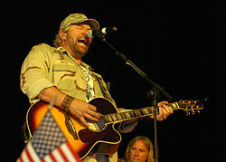 A man in a camouflage jacket and cap playing a guitar and singing into a microphone