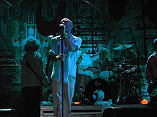 A blue-tinted photograph of musicians in front of an industrial background. From left to right: a long-haired male stands with his back to the camera playing bass guitar, a middle-aged Caucasian male sings into a microphone, a middle-aged Caucasian male plays behind a black-and-silver drum set on a riser, and a guitar player is mostly cropped from the extreme left of the photo.