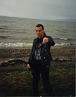 A man, Chuck Schuldiner, is shown on a dark shoreline. He has long hair, black pants and a black shirt, and a black leather jacket.