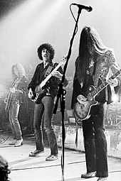 Three members of the band Thin Lizzy are shown onstage. From left to right are a guitarist, bass player, and another electric guitarist. Both electric guitarists have long hair.